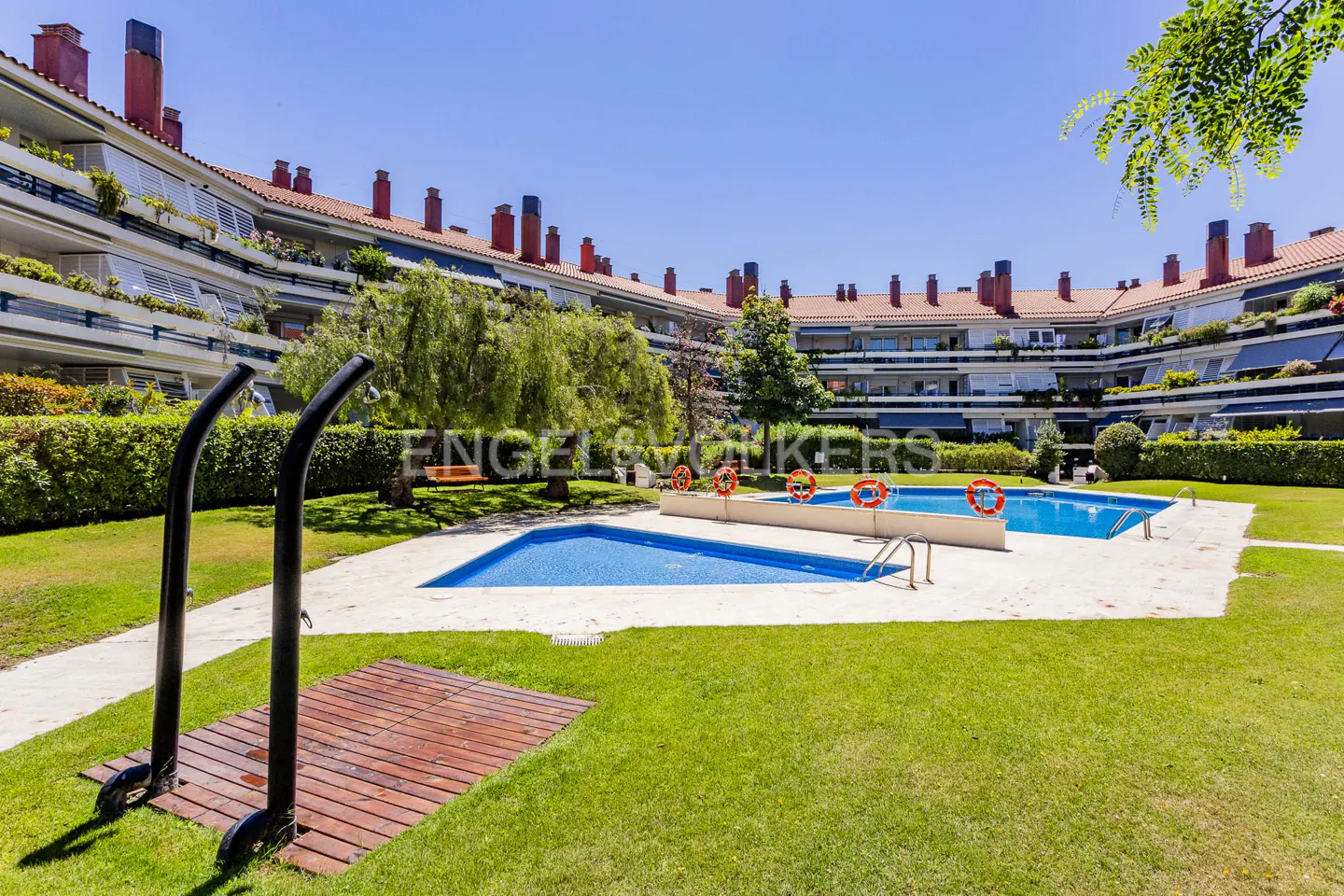 Outdoor pool area with blue water, surrounded by green grass, trees, and a multi-story building with red tile roofs. Showers in foreground.