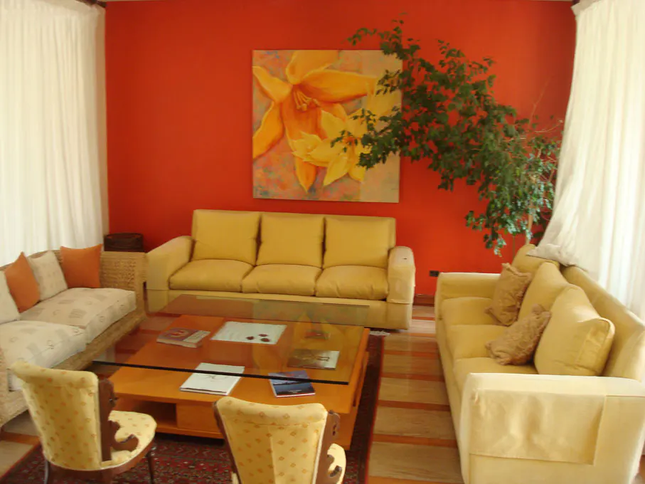 Living room with orange walls, yellow sofas, and a glass-topped coffee table. A painting of a yellow flower hangs above the sofa. Curtains are white.