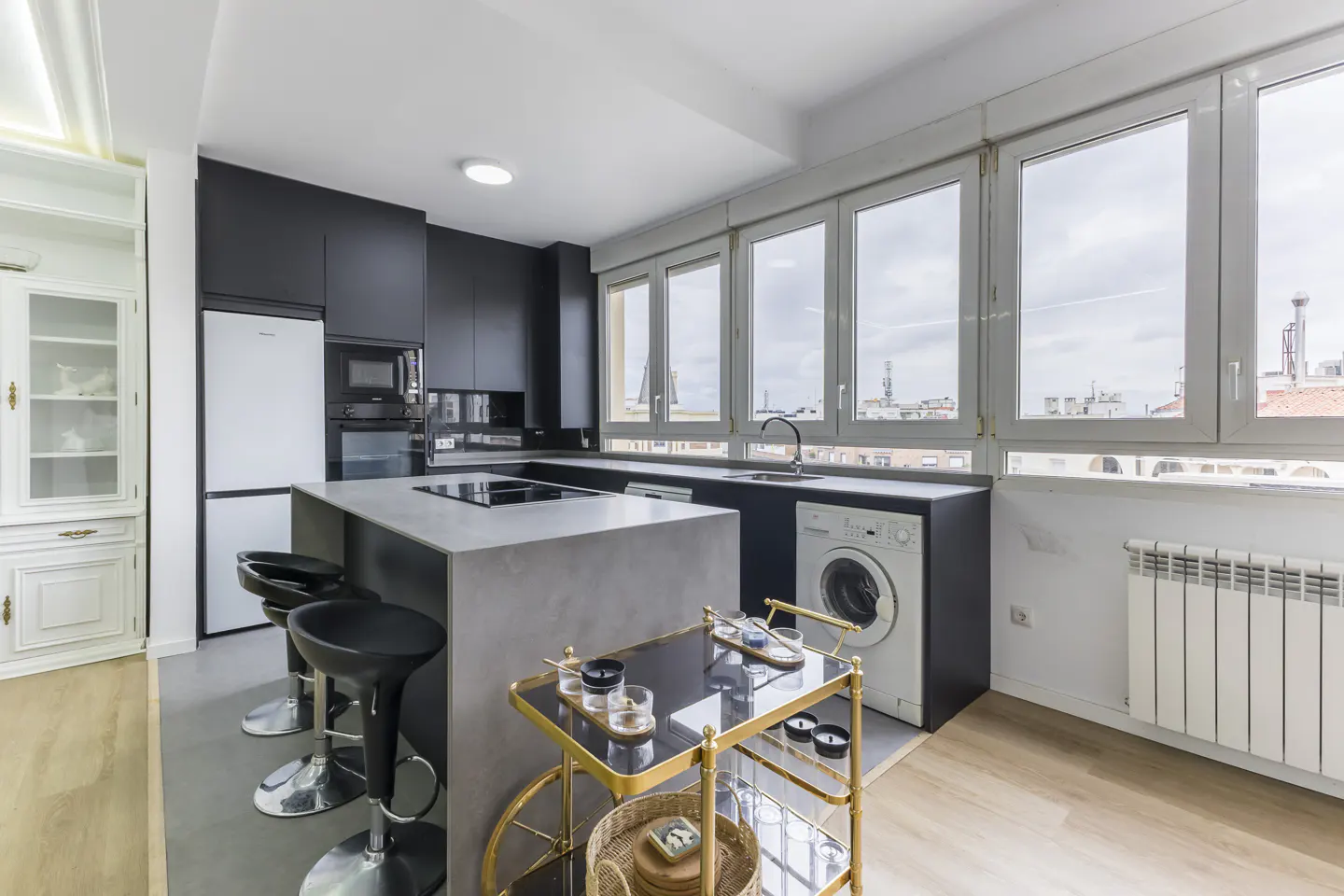Modern kitchen with black cabinets, a gray island with black stools, and a gold bar cart. Large windows offer a city view.
