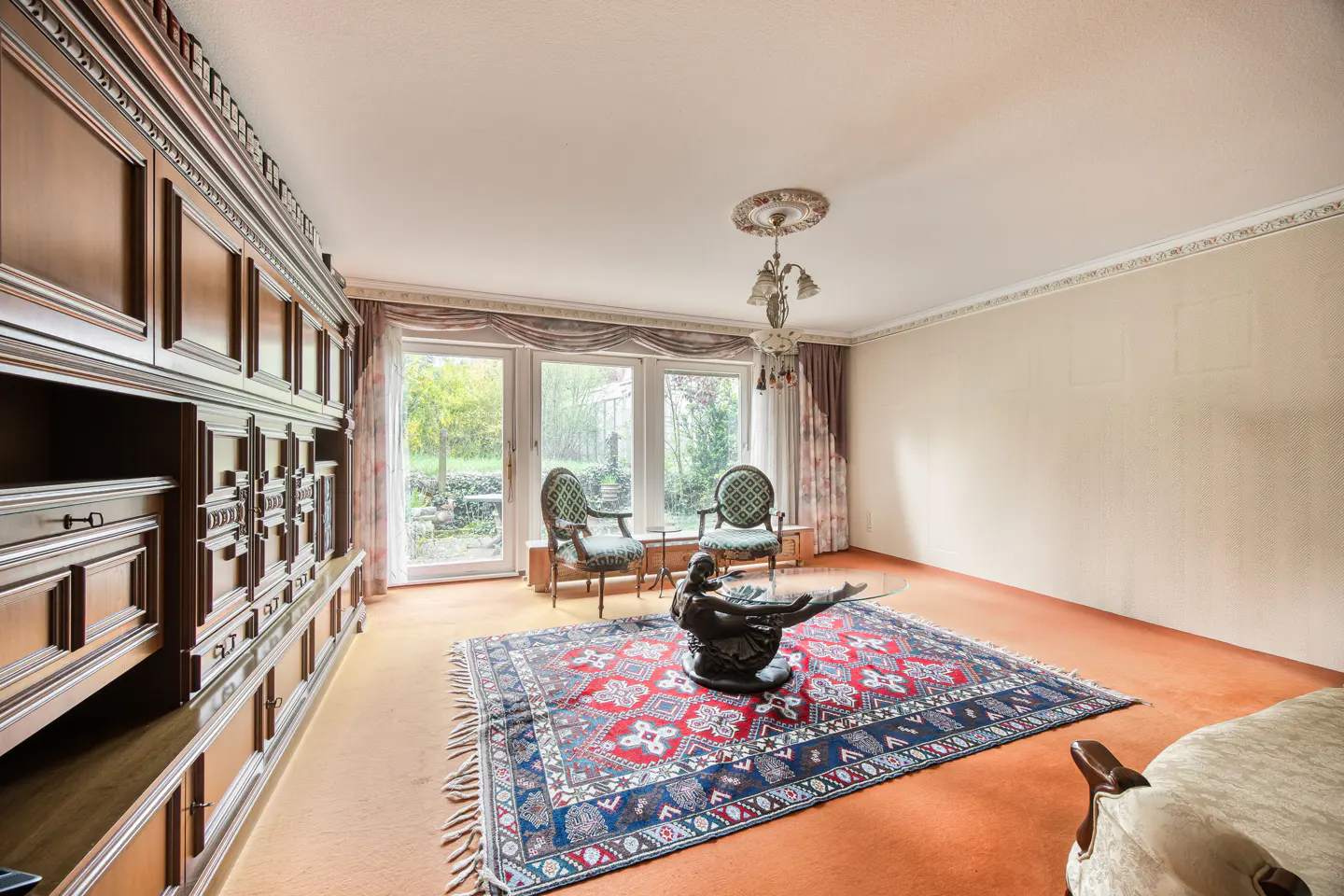 Living room with a large wooden cabinet, two chairs, a patterned rug, and a sculpture on a glass table.