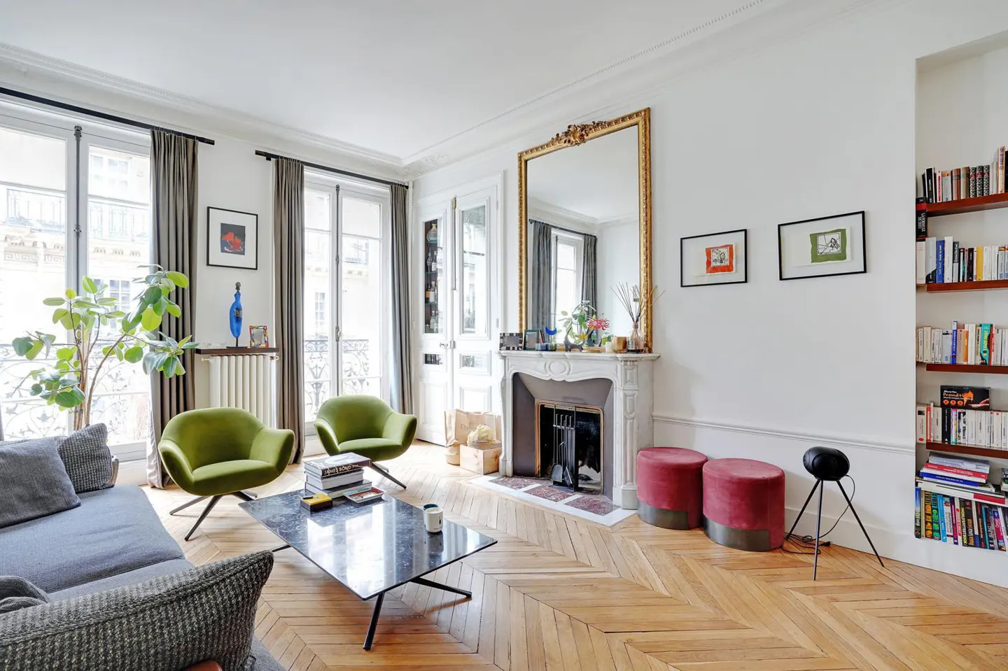 Bright living room with herringbone wood floors, white walls, and tall windows. Green chairs, a gray sofa, and a marble fireplace add color.