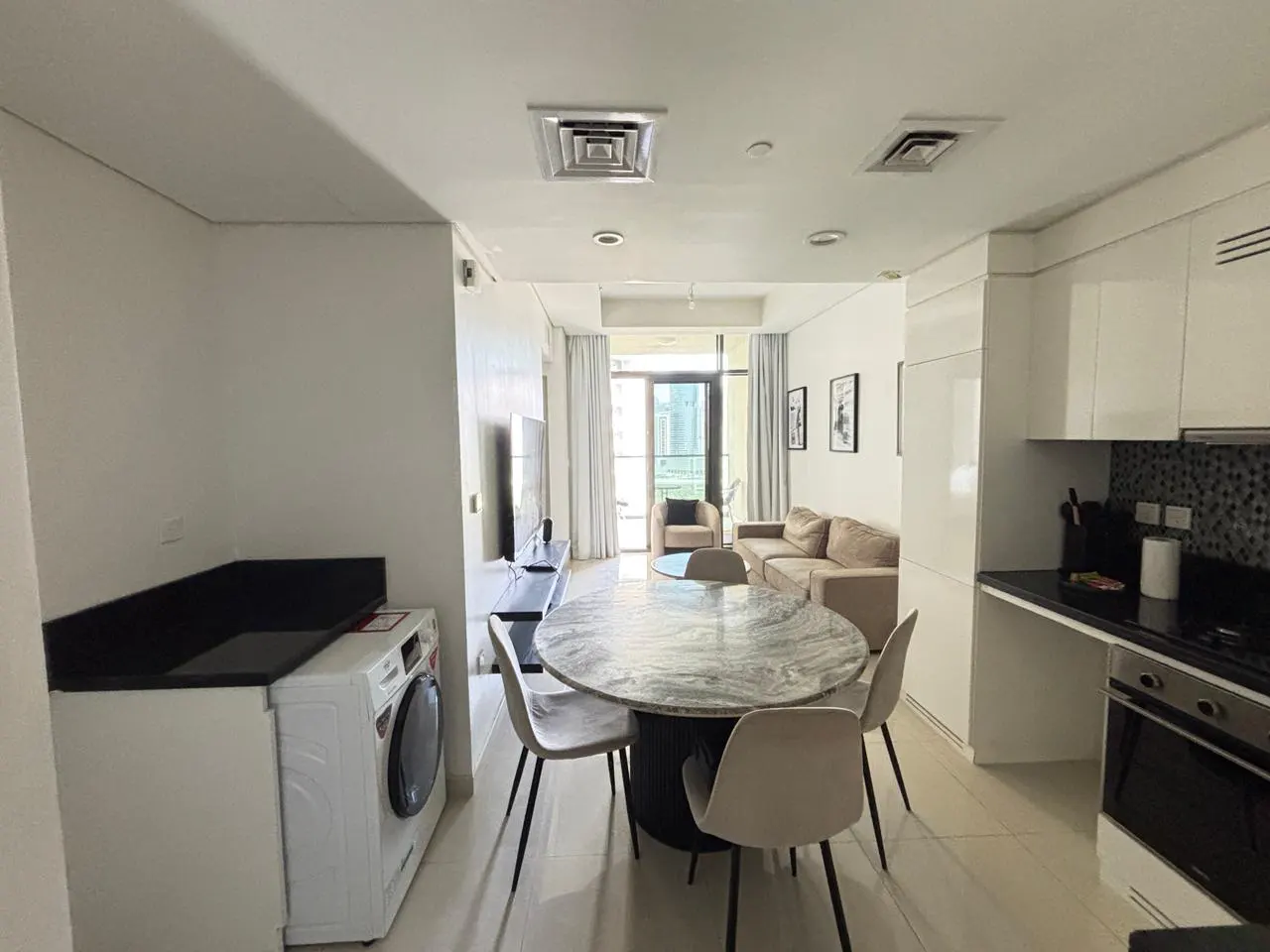 A bright, modern apartment interior with a kitchen, dining area, and living room. A washing machine is visible near a marble table with four chairs.