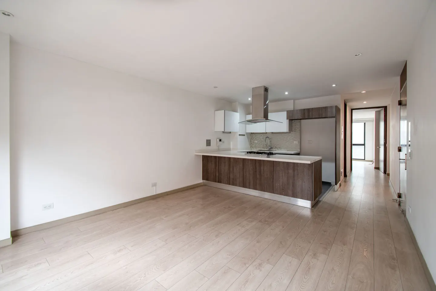 Bright, empty apartment with light wood floors, white walls, and an open kitchen with brown cabinets and stainless steel appliances.