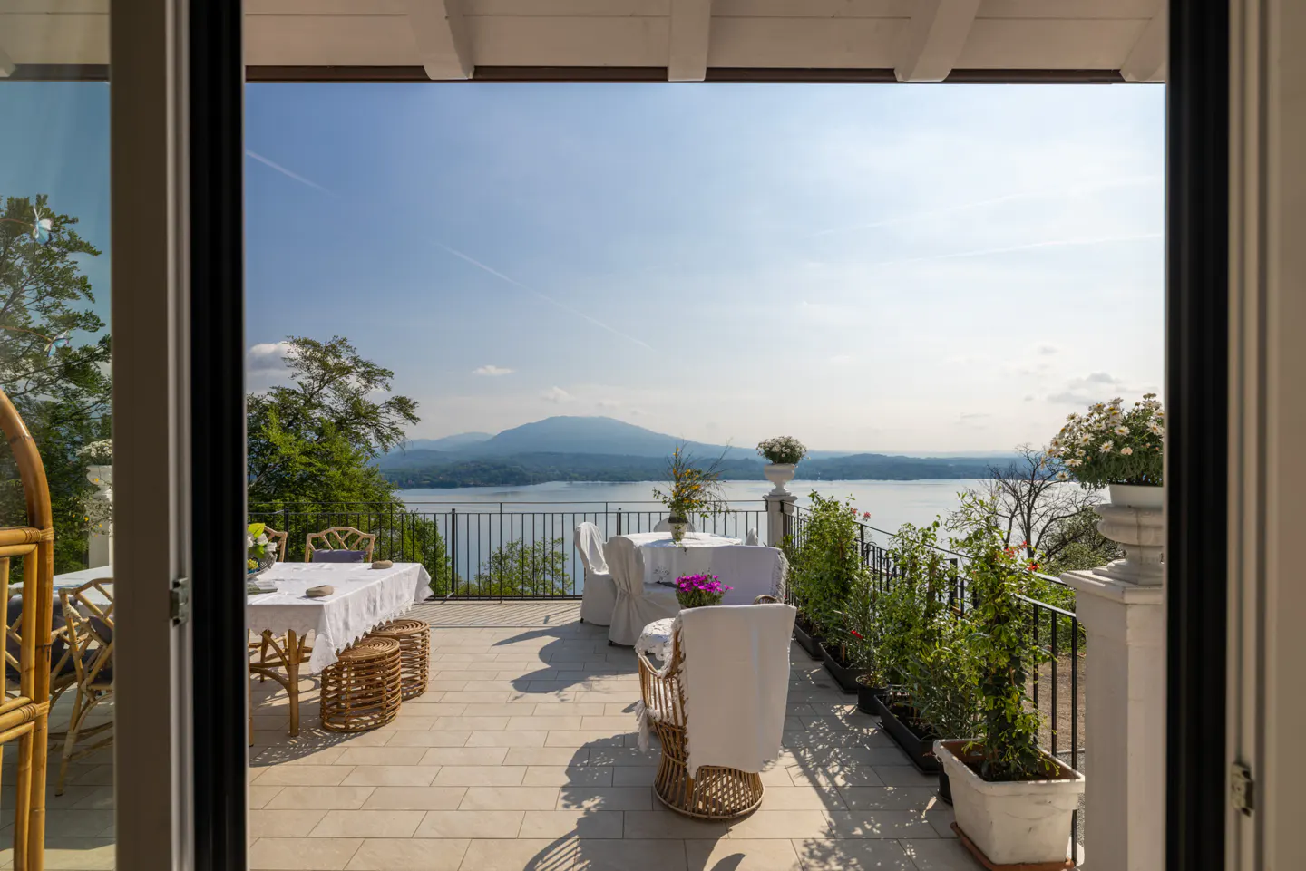 View from open doorway onto a patio with tables, chairs, and a lake view. Tables are covered with white tablecloths. The sky is blue.