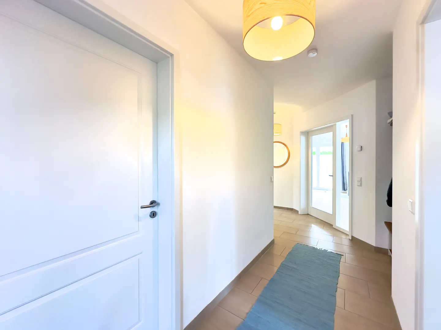 Bright hallway with white walls, a blue rug on the tiled floor, and a white door on the left. A light fixture hangs from the ceiling.