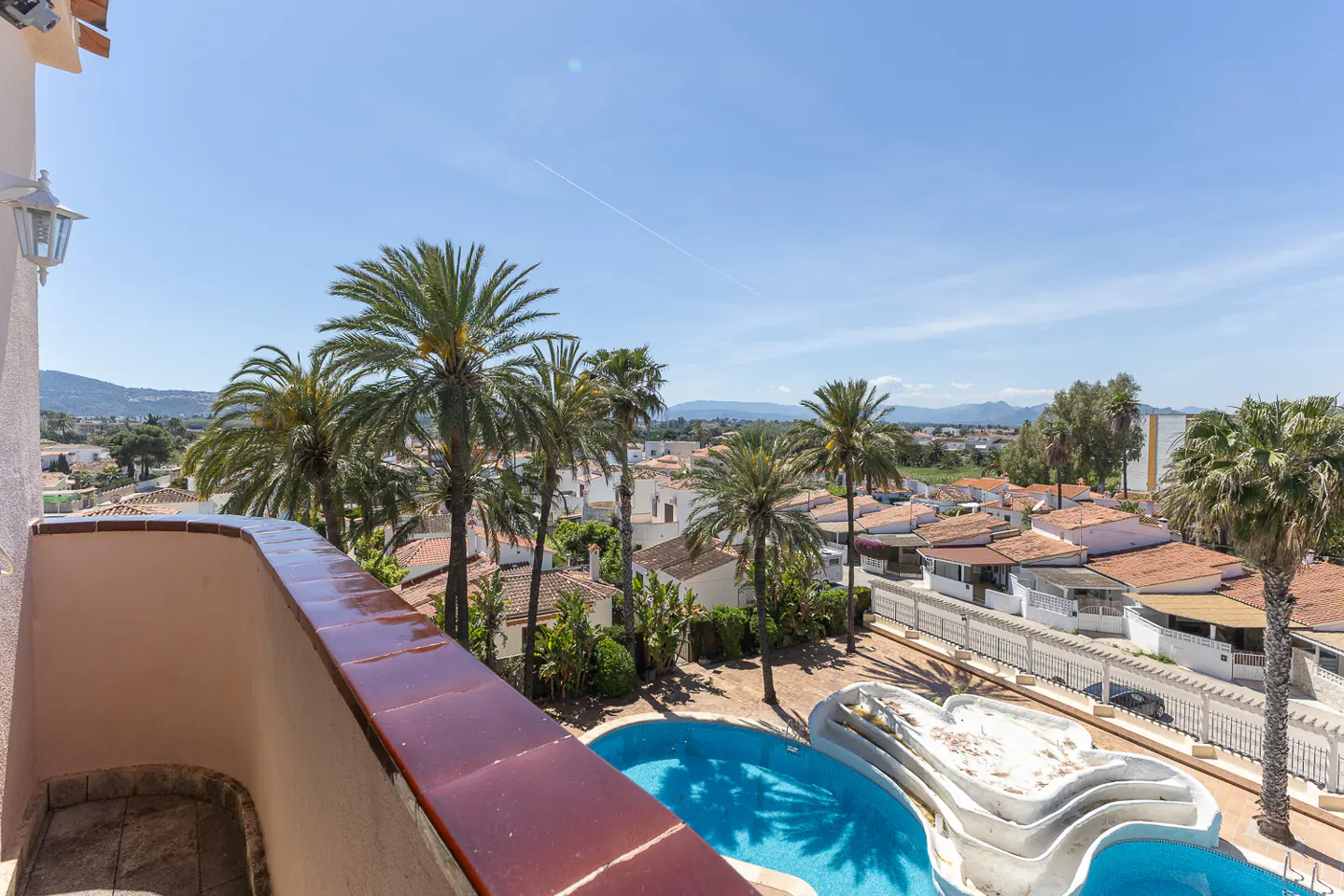 View from balcony overlooking a pool, palm trees, and houses with red tile roofs under a blue sky.