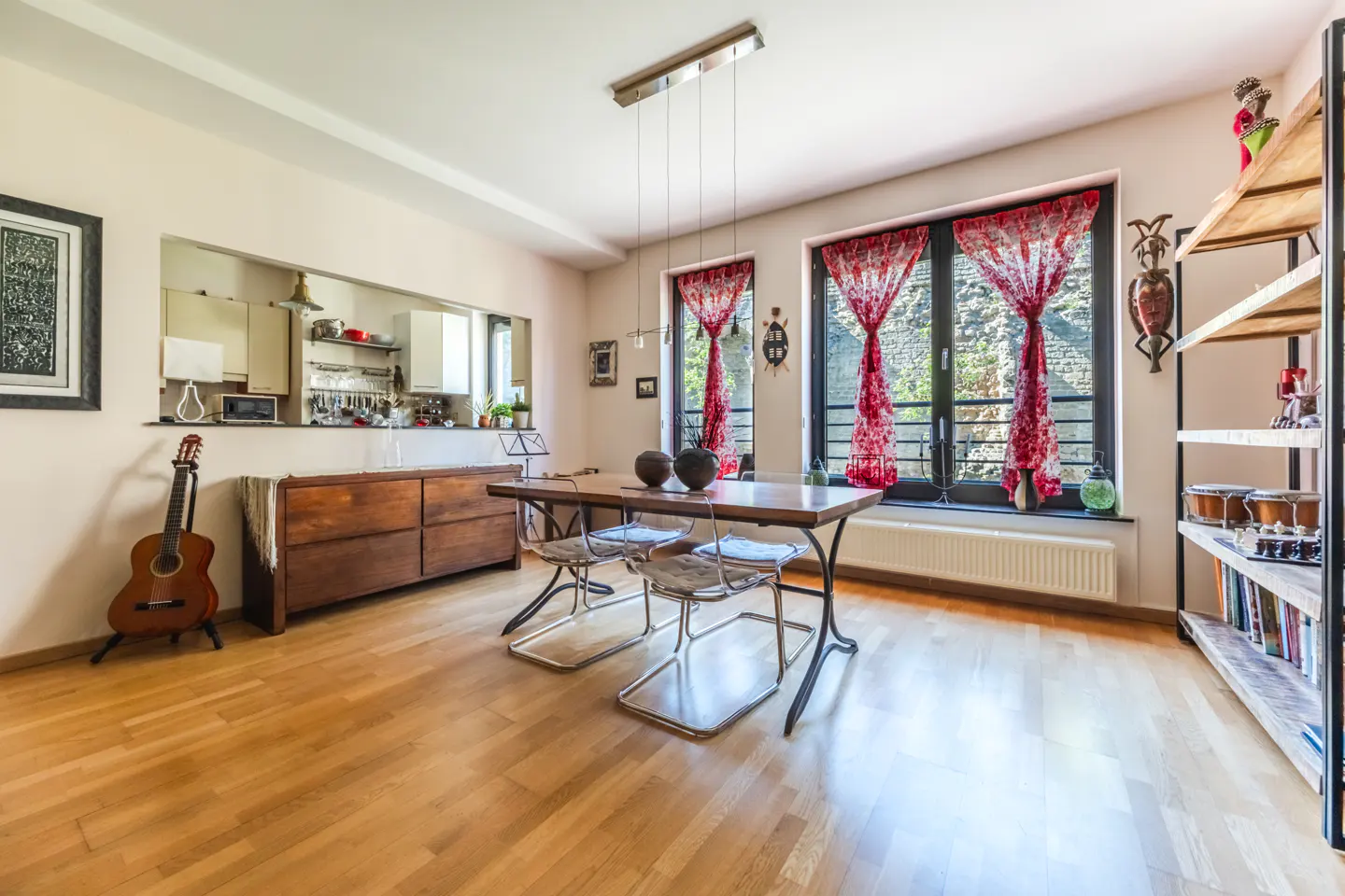 A bright dining room with wood floors, a table with four chairs, and red patterned curtains on the windows. A guitar leans against a cabinet.