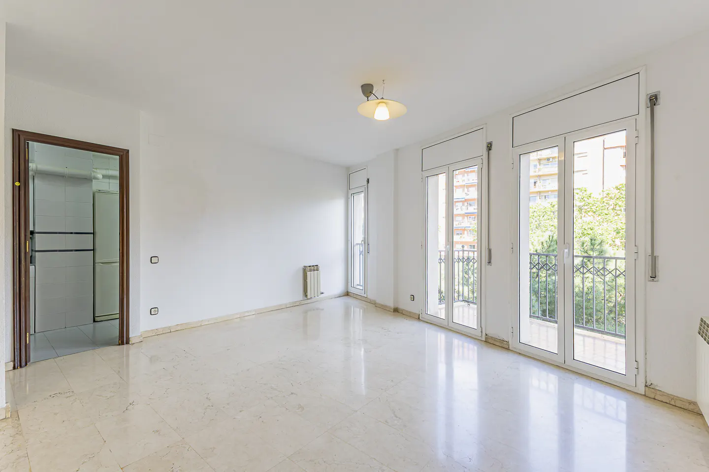 Bright, empty room with marble floors, white walls, and a brown door. Balconies with iron railings offer views of trees and a building.