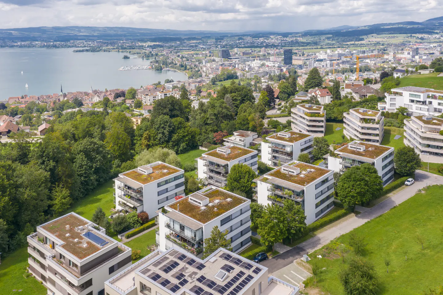 Aerial view of modern white apartments with green roofs, overlooking a lake and city skyline.