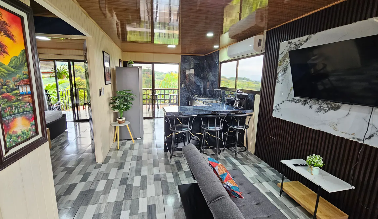 A modern living space with a kitchen island, gray sofa, and a view of lush greenery.
