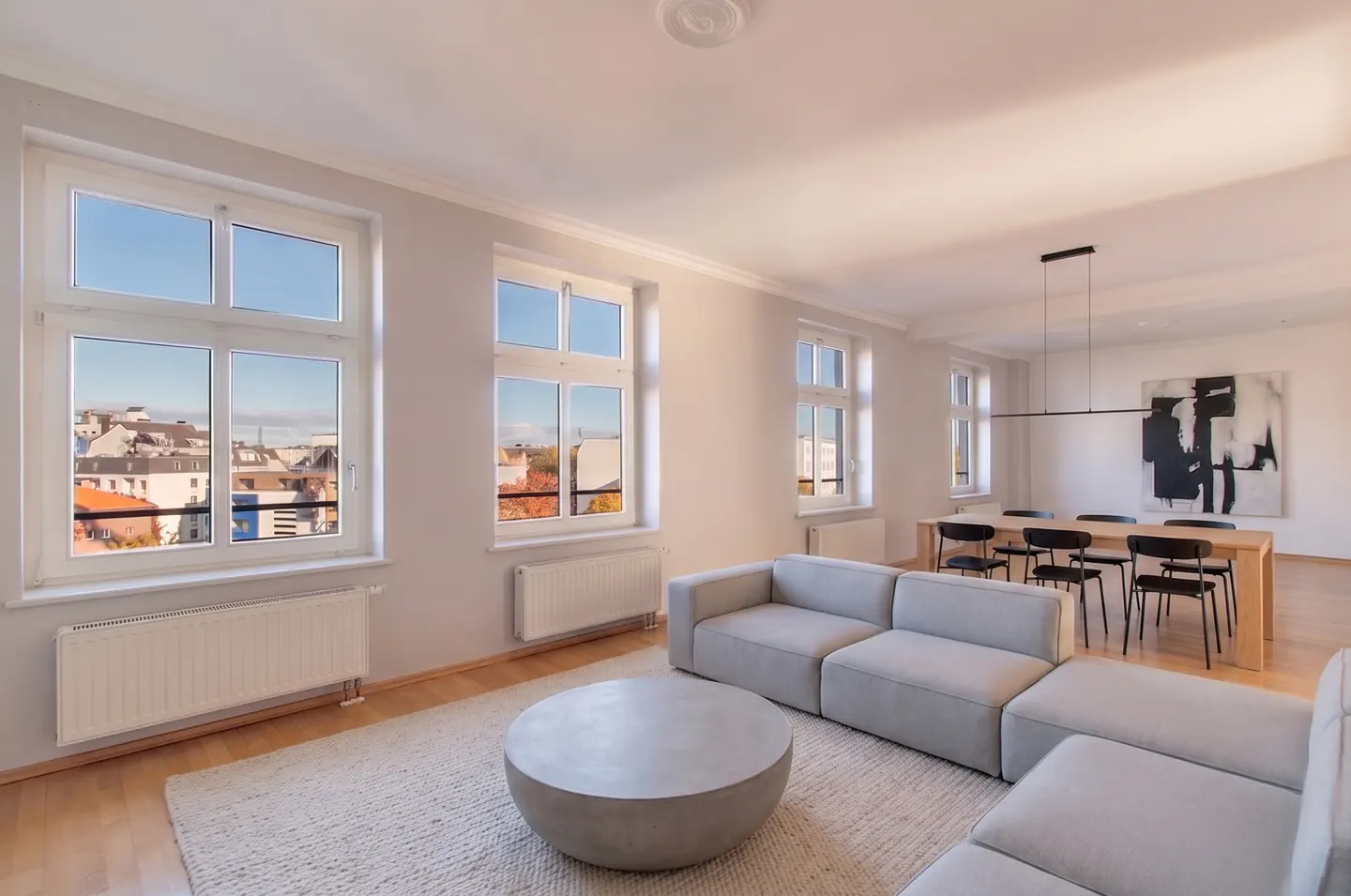Bright living room with a gray sectional sofa, round coffee table, and dining table with black chairs. Large windows offer a city view.