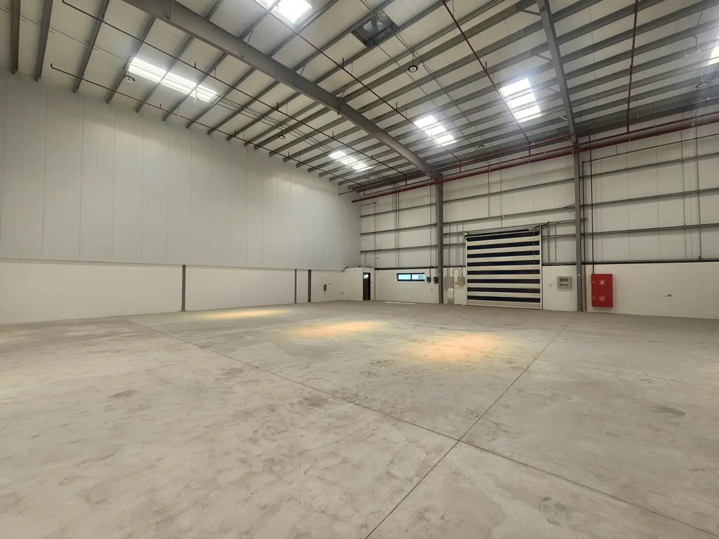 A large, empty warehouse with a concrete floor, white walls, and a high, exposed ceiling with metal beams. A large, striped door is on the right.