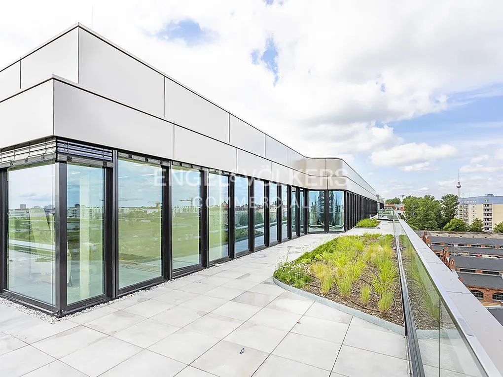 Rooftop view of a modern building with large windows, a tiled patio, and greenery. The city skyline is visible in the background.