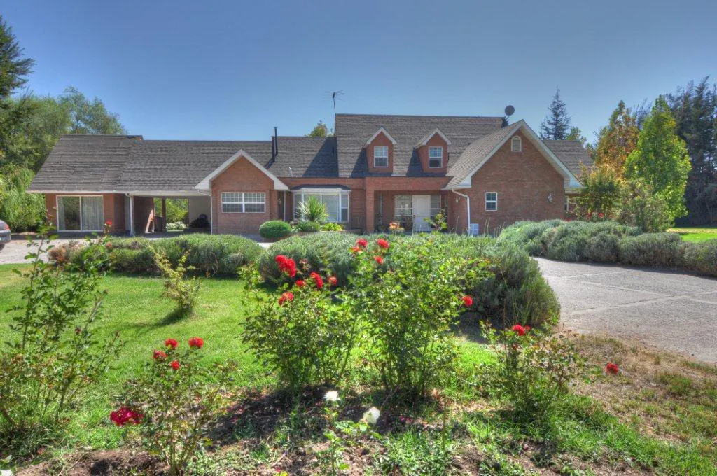Brick house with a dark roof, dormer windows, and a covered carport. Red roses and green bushes line the front yard.