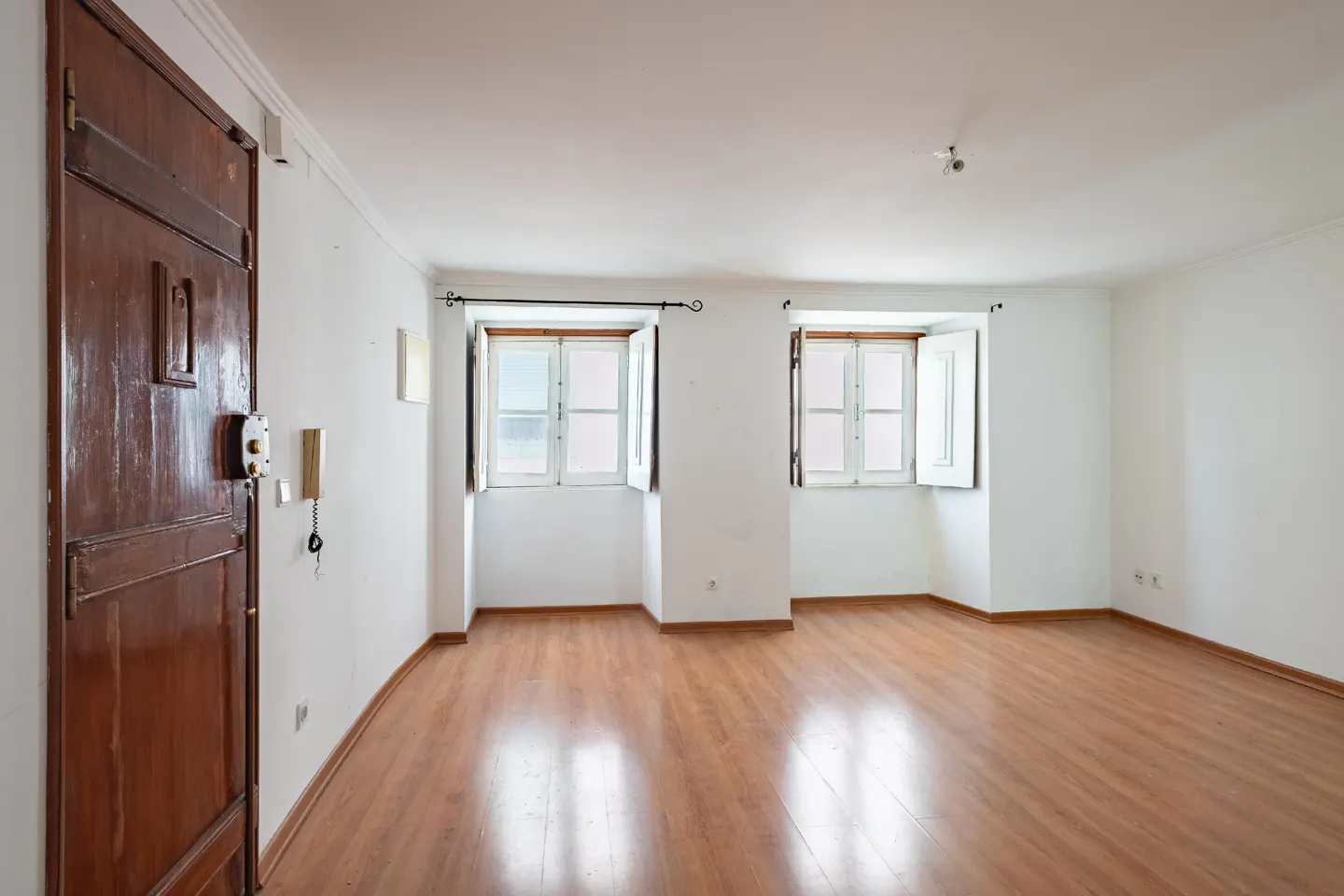 Bright, empty room with wood floors, white walls, and two windows with open white shutters. A dark wood door is on the left.