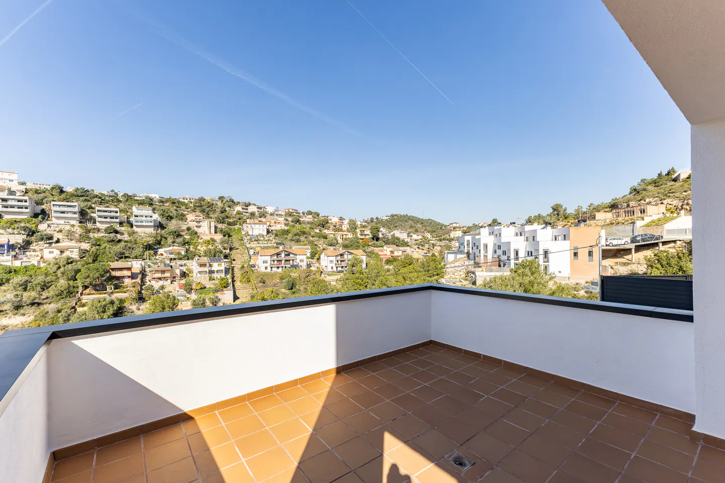 Balcony with brown tile floor and white walls overlooking a hillside neighborhood under a clear blue sky.