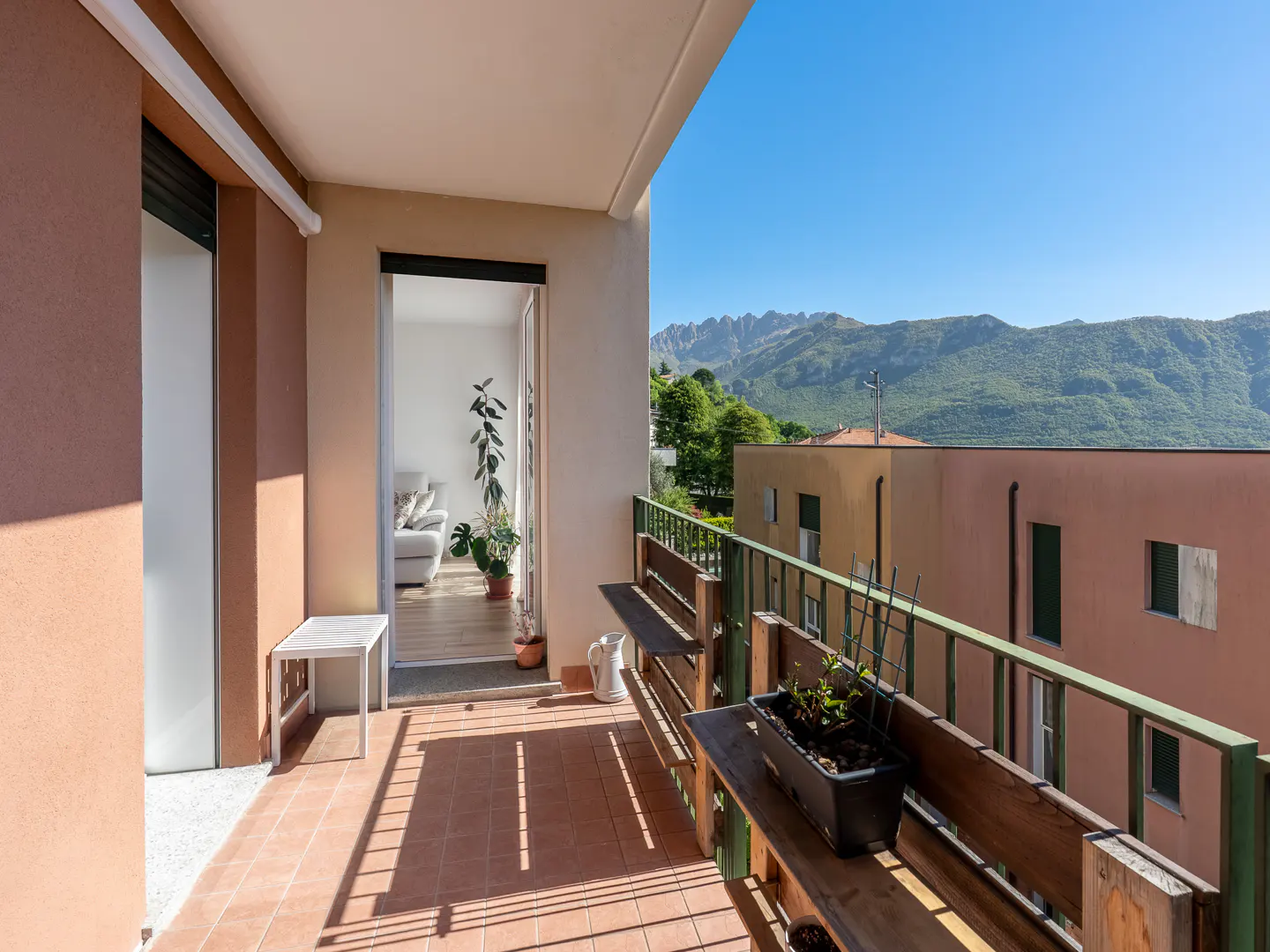 Balcony with terracotta tiles, wooden benches, and a view of mountains and buildings under a clear blue sky. Interior visible through open door.