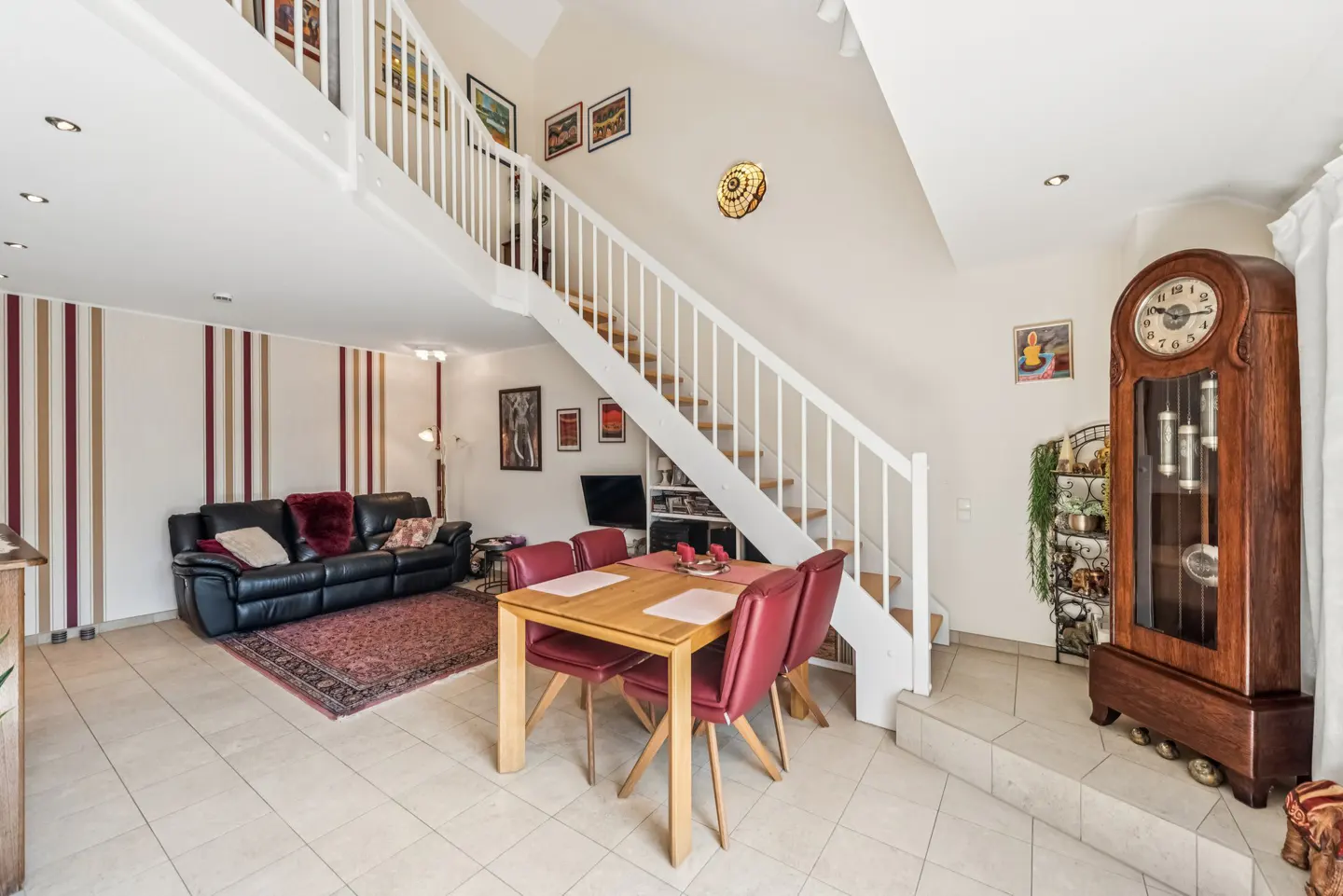 Open-concept living room with a black leather sofa, wooden dining table with red chairs, and a white staircase leading to a loft.