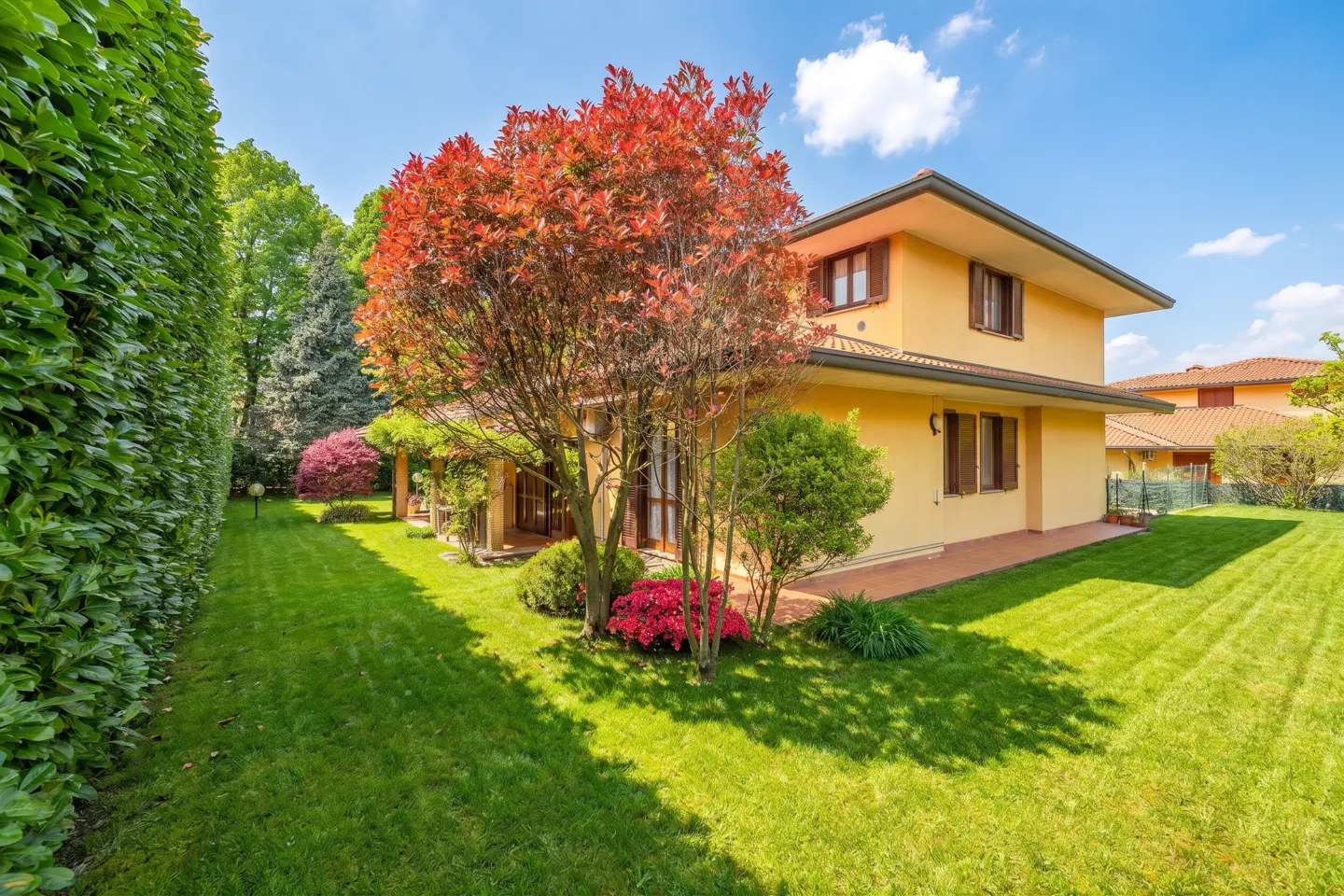 Two-story yellow house with brown shutters, a red-leafed tree, and a green lawn under a blue sky.