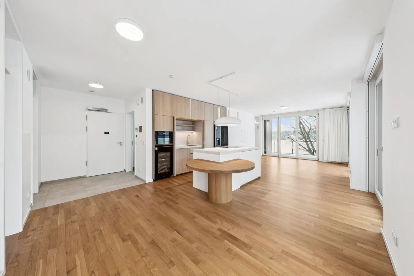 Modern open-plan kitchen with light wood cabinets, a white island, and a circular wooden dining table on hardwood floors.