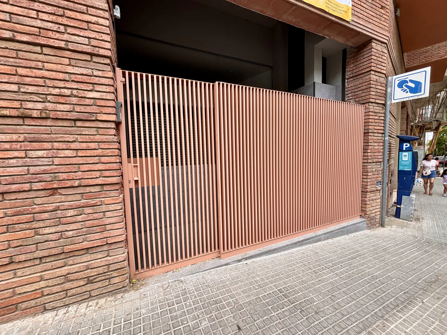 Exterior shot of a building with a brick facade and a salmon-colored metal gate. A parking meter and handicap sign are visible on the sidewalk.