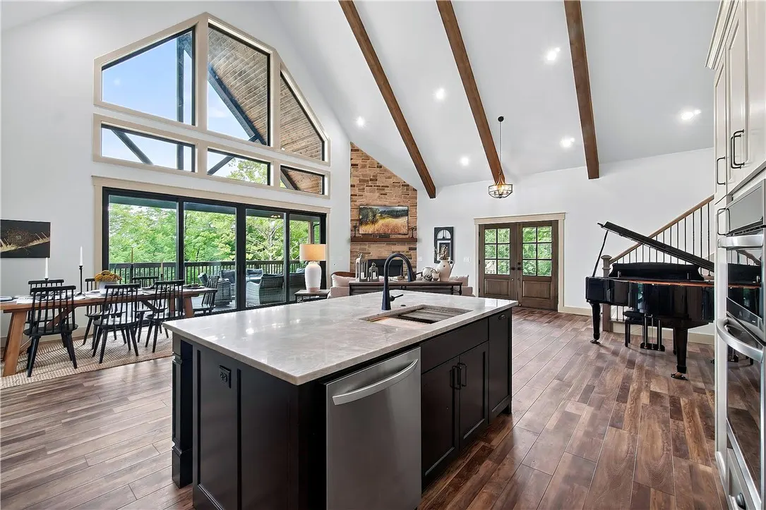 Modern open-concept kitchen with dark island, wood floors, vaulted ceiling with beams, and a grand piano in the living area.