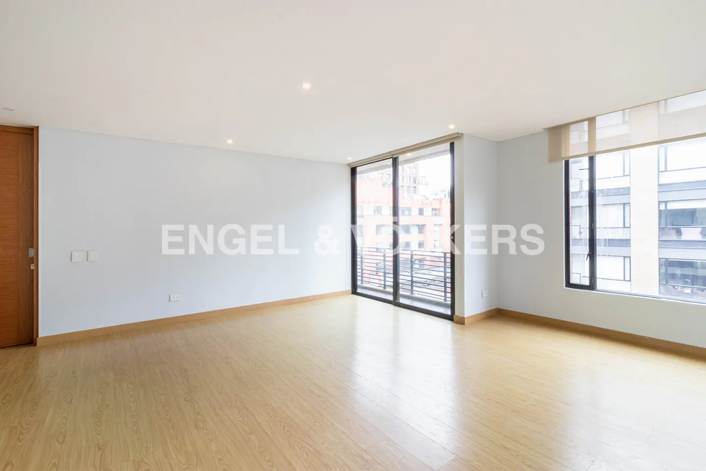Empty room with light wood floors, pale blue walls, and large windows overlooking a city building.