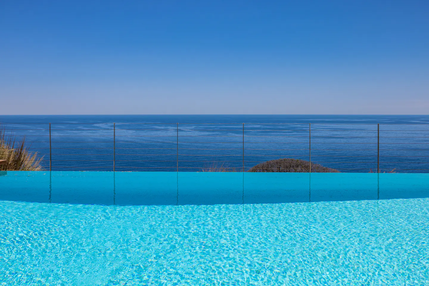 Infinity pool with turquoise water merges into the blue ocean under a clear sky. A glass fence separates the pool from the sea.