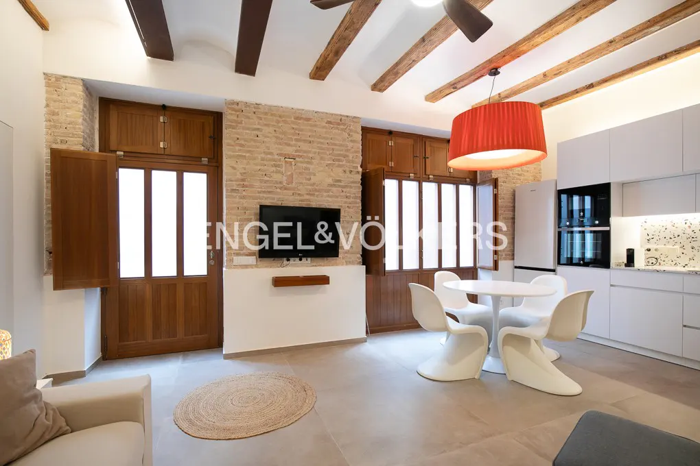 A living room with exposed brick, wood beams, and a modern kitchen. A round table with white chairs sits under a red pendant light.