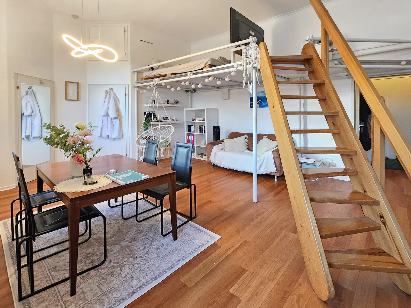 Modern studio apartment with a dining table, loft bed, wooden staircase, hanging chair, and a sofa on hardwood floors.