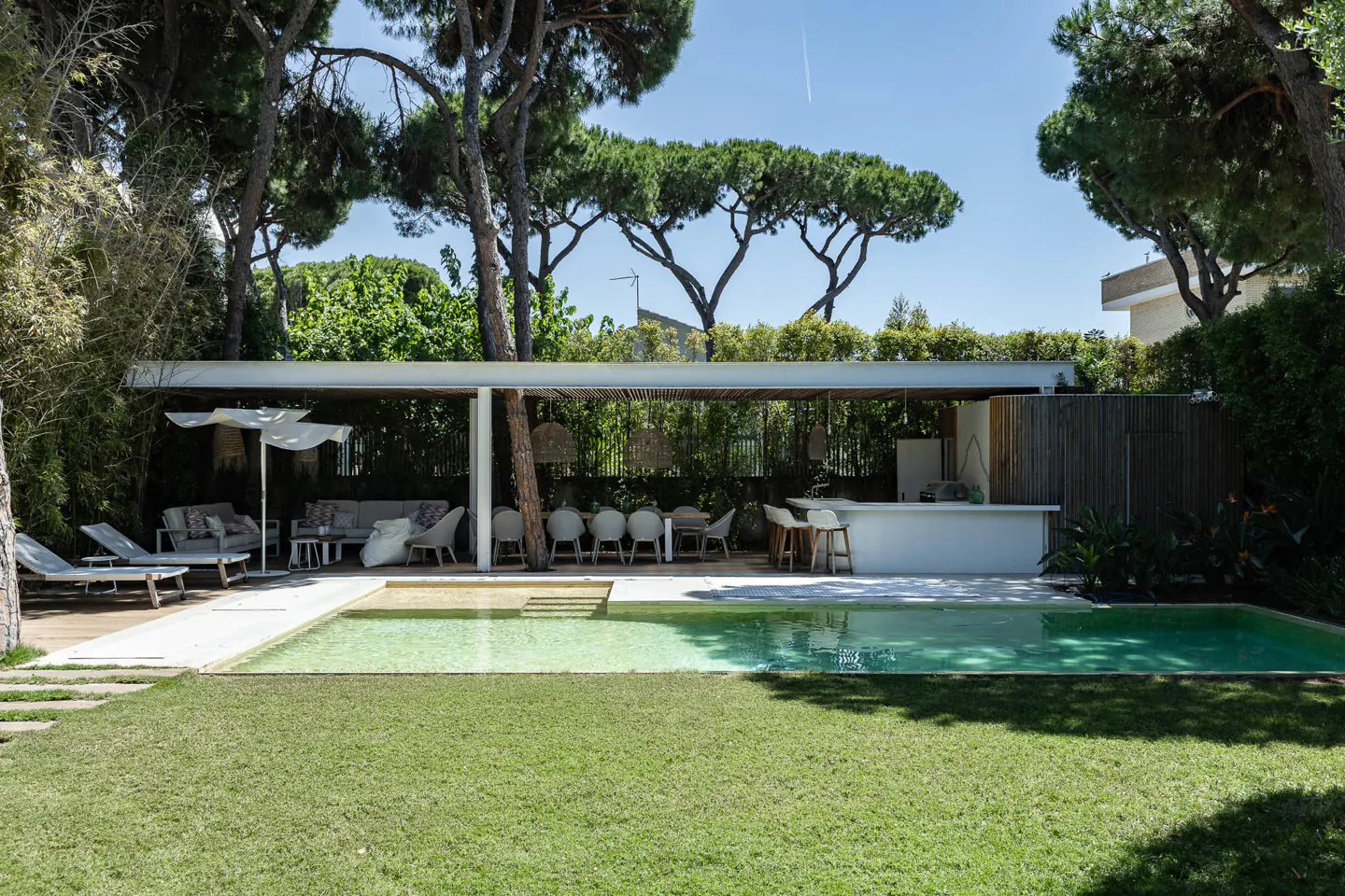 A backyard with a green lawn, a pool, and an outdoor living area with a white bar and seating.
