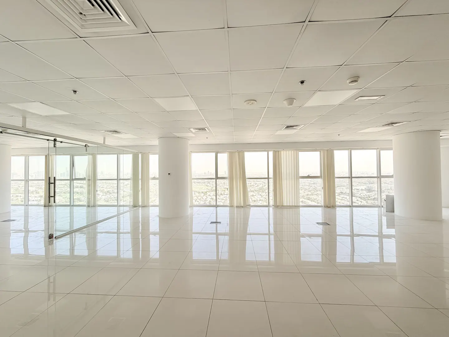 An empty, bright office space with white tile floors, large windows, and white support columns.
