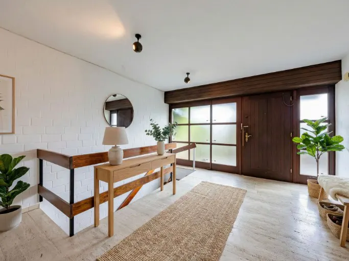 Bright entryway with a wood door, frosted glass panels, and a long jute rug. A console table with a lamp and plants sits near a wooden railing.
