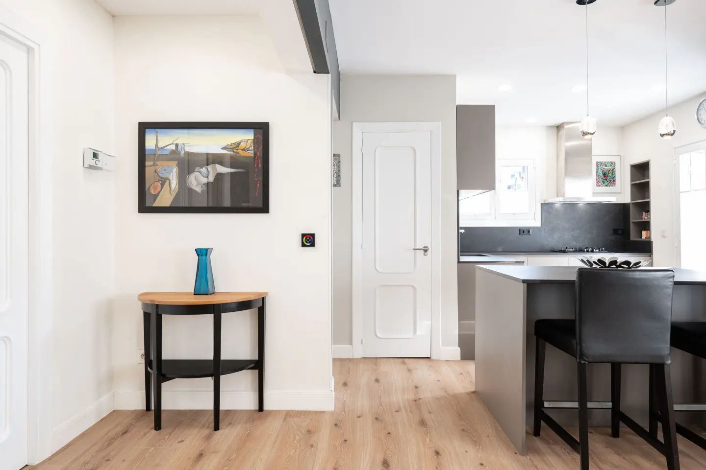 Bright, modern kitchen with light wood floors. A black console table holds a blue vase under a framed painting. A gray island has black stools.