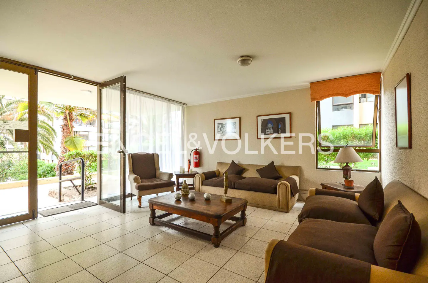 Living room with brown sofas, a wooden coffee table, and an open door to a patio with palm trees.
