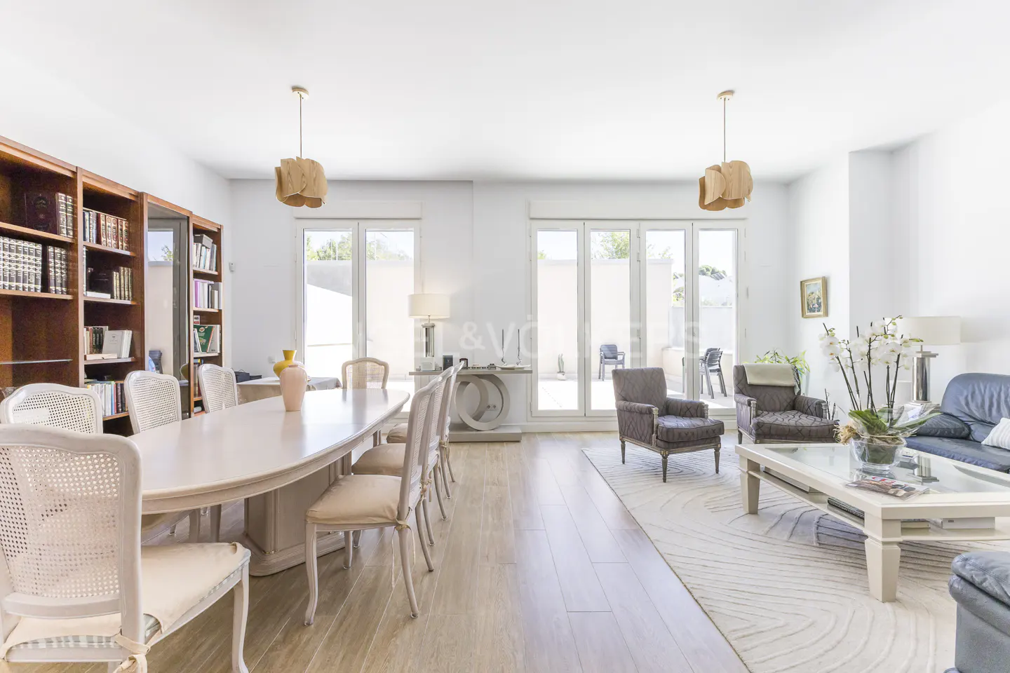 Bright, open-plan living and dining area with a long table, chairs, and a bookshelf. Two armchairs and a sofa are in the living area.
