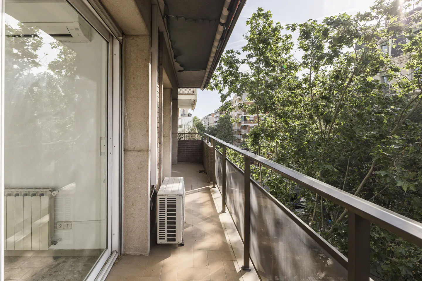 A sunlit balcony with a metal railing overlooks lush green trees and a distant building. An air conditioning unit sits on the tiled floor.