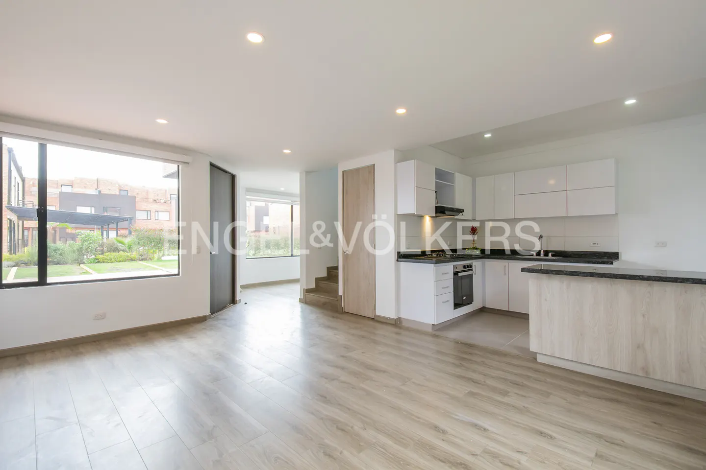 Bright, open-concept living space with wood floors, white kitchen cabinets, and a large window overlooking a green lawn.