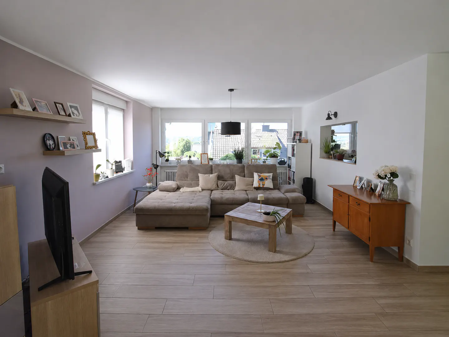 Bright living room with a large beige sectional sofa, wooden coffee table, TV, and a wooden sideboard on light wood floors.
