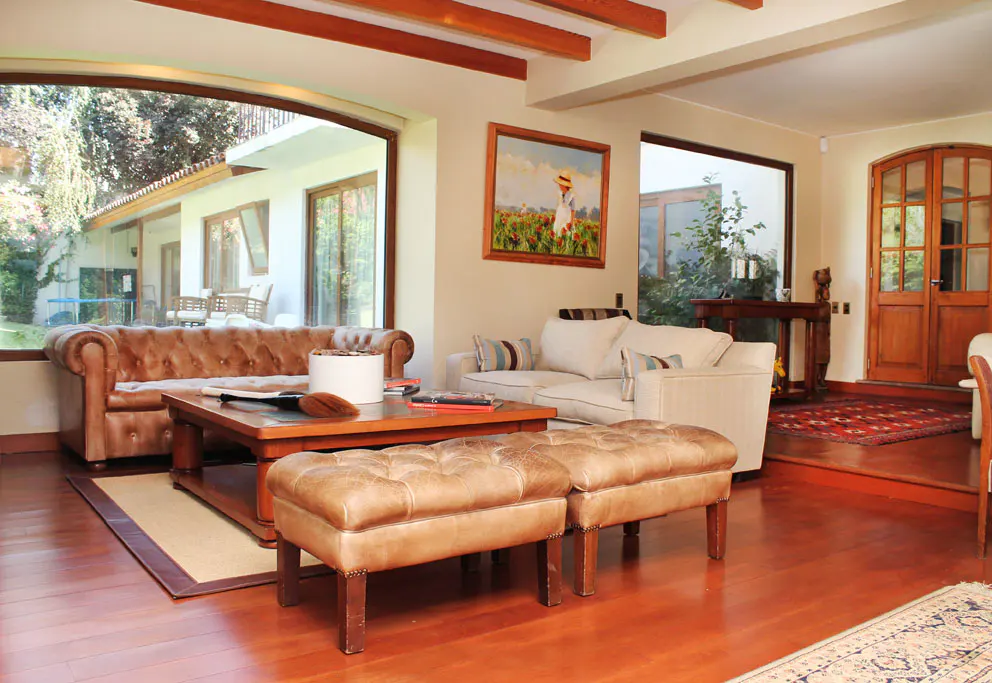 Living room with hardwood floors, brown leather sofa, white sofa, and two leather ottomans. Large windows overlook a green yard.