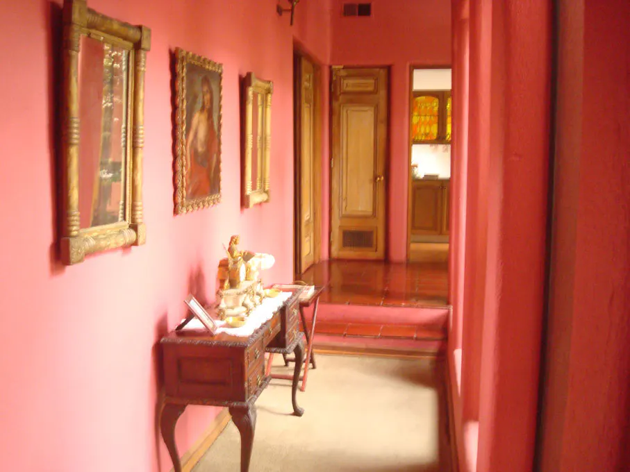 Hallway with pink walls, wooden floors, and antique furniture. Paintings hang above a dark wood table with decorative objects.