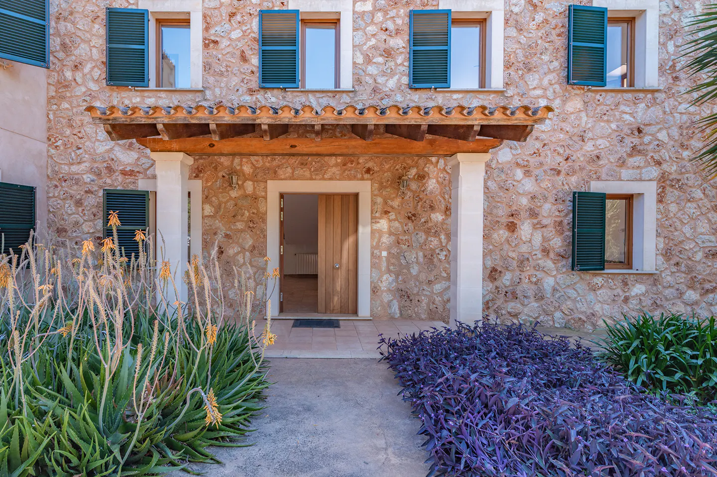 Stone house facade with a wooden porch, open front door, green shutters, and a garden with aloe and purple plants.