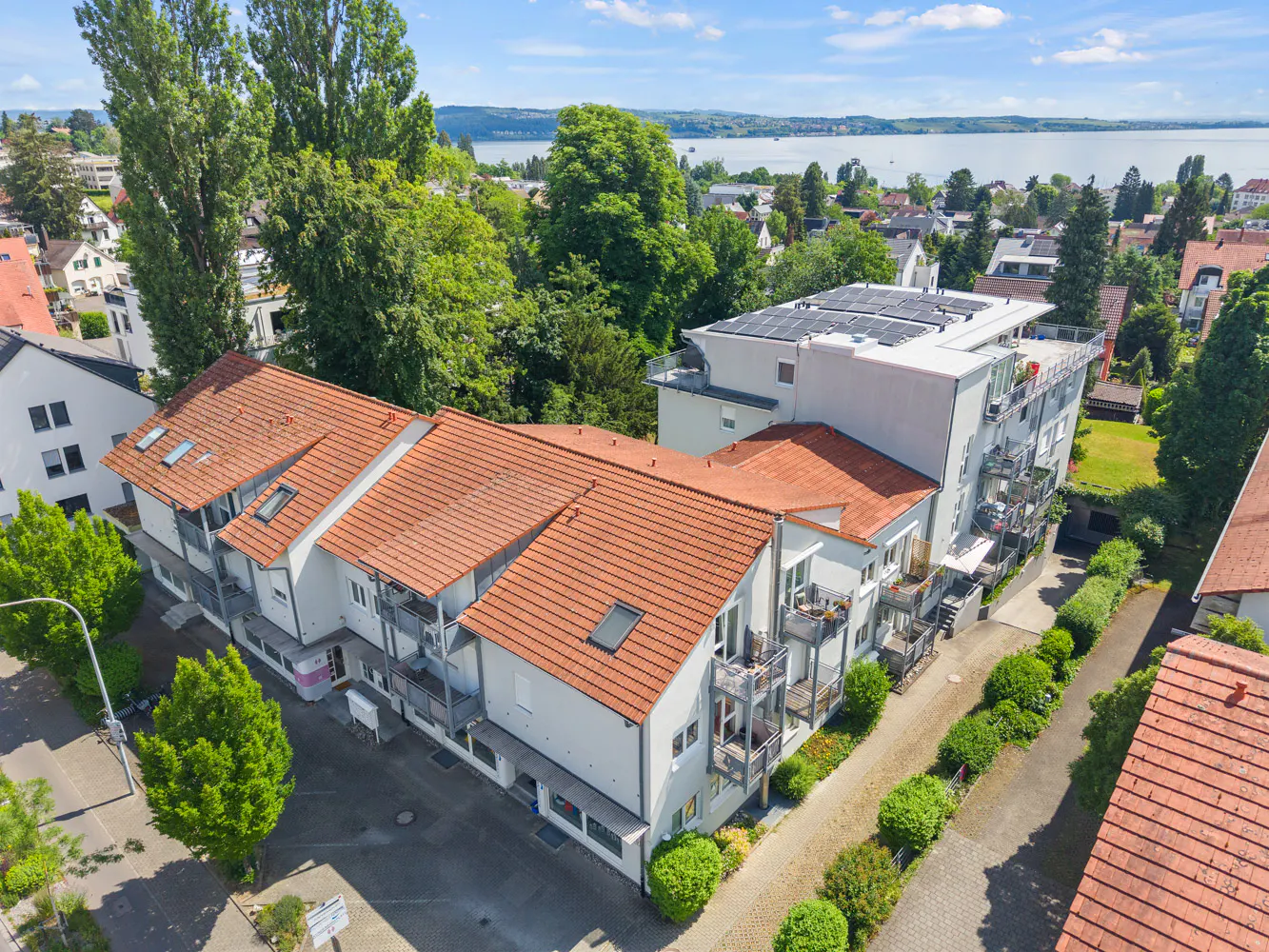 Aerial view of a white apartment complex with red tiled roofs, balconies, and solar panels, set near a lake and greenery.