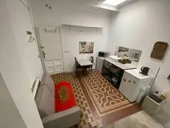 Small studio apartment with a grey sofa, dining table, kitchenette with white cabinets, and patterned tile flooring.