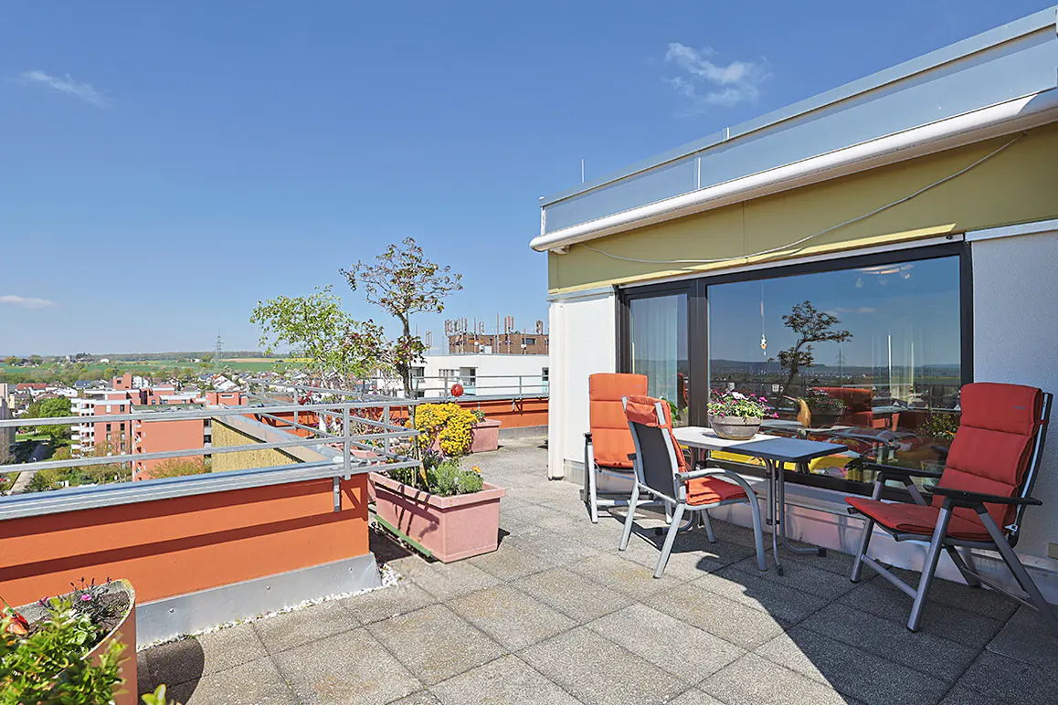 A rooftop patio with red chairs and a table overlooks a cityscape under a blue sky. Potted plants add greenery to the outdoor space.