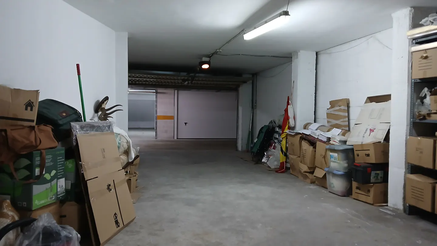 A concrete storage garage with white walls, fluorescent lighting, and stacks of cardboard boxes lining both sides.