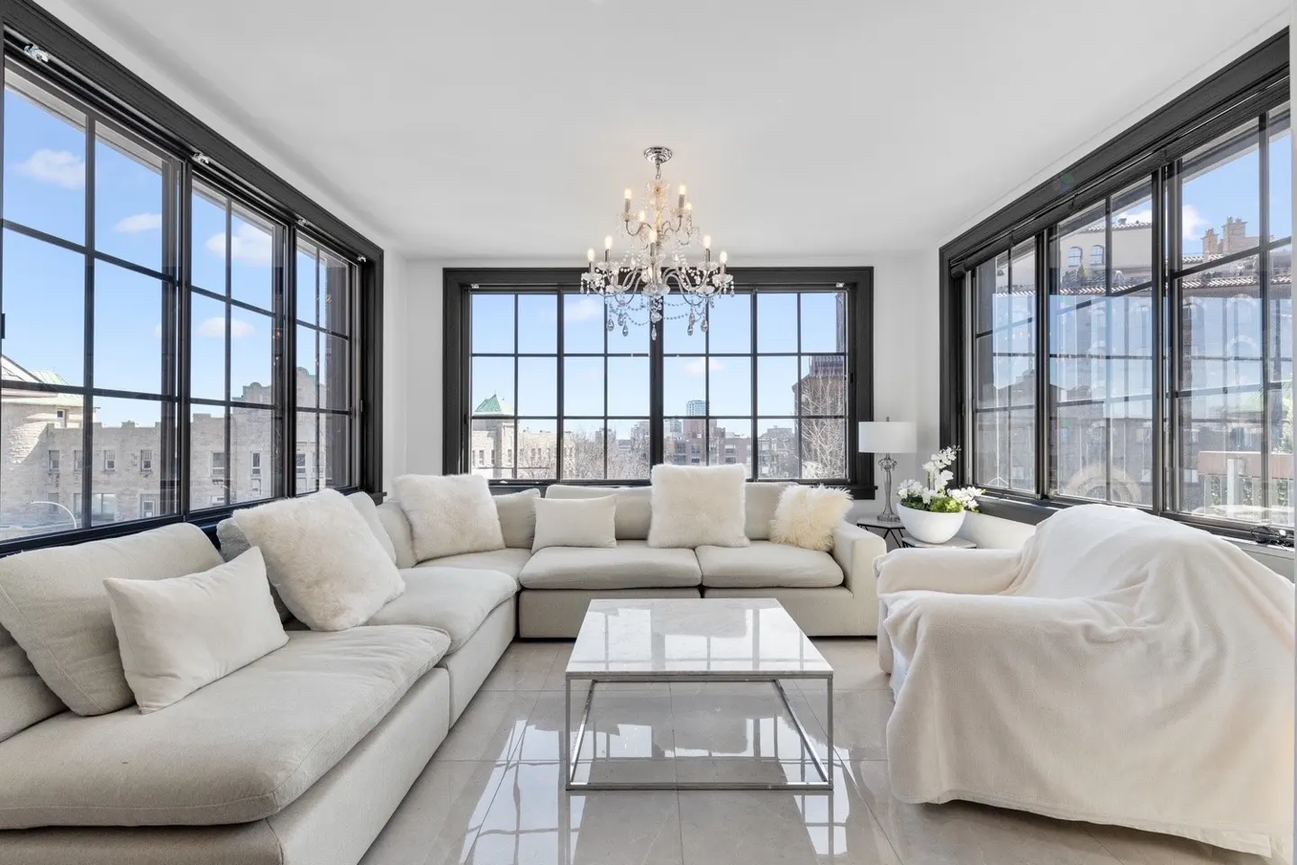 Bright living room with white sectional sofa, marble coffee table, and large windows with black frames overlooking a city.