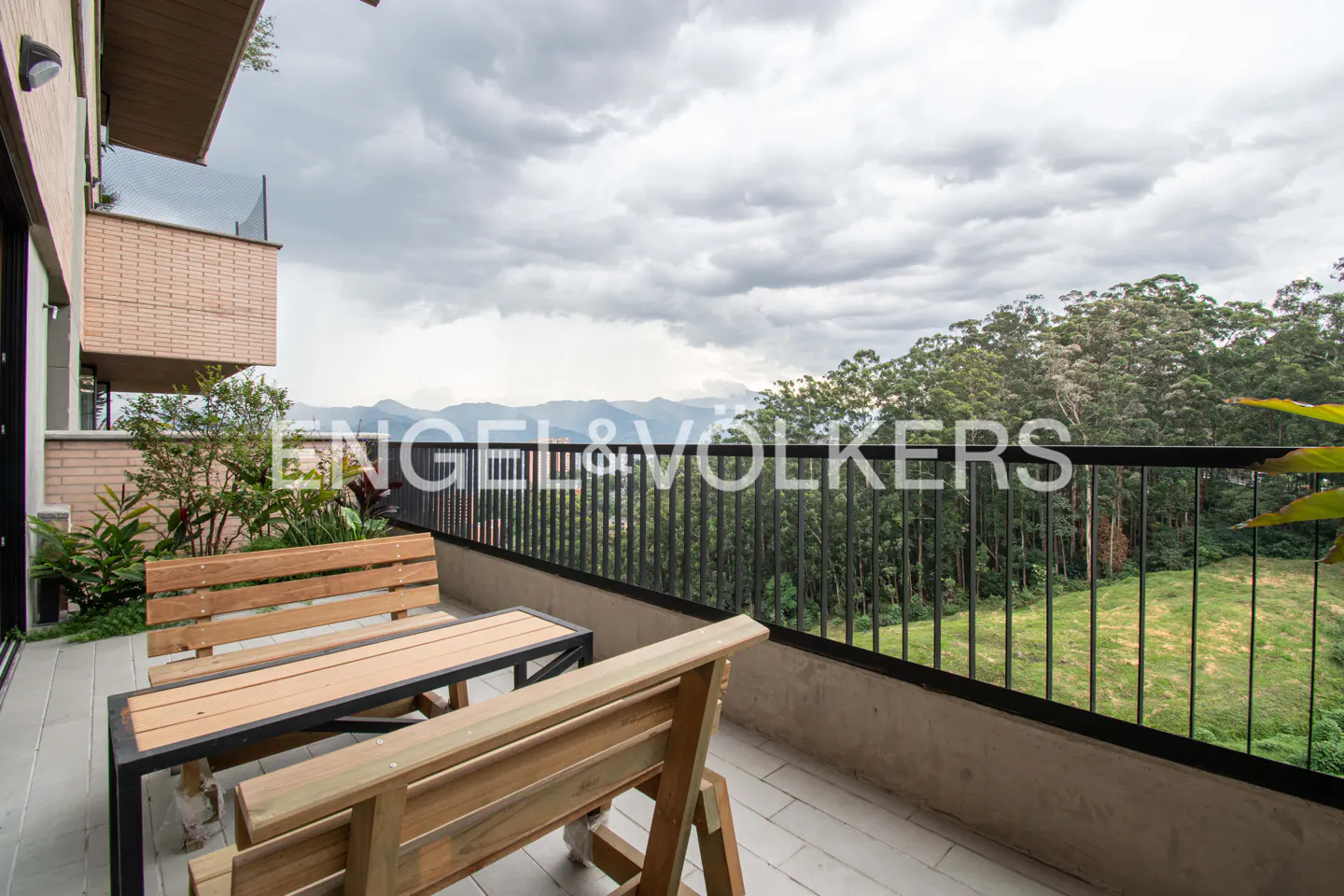 Balcony view with wooden bench and table, black railing, and lush green trees under a cloudy sky.