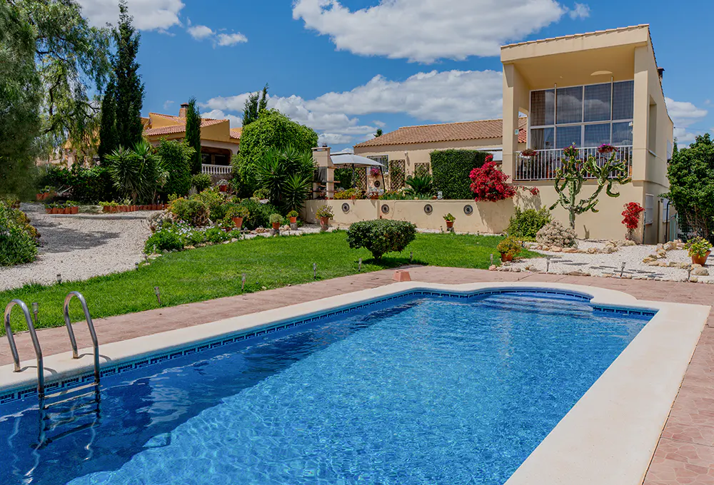 A sunny backyard with a blue tiled pool, green lawn, and a tan house with a balcony.