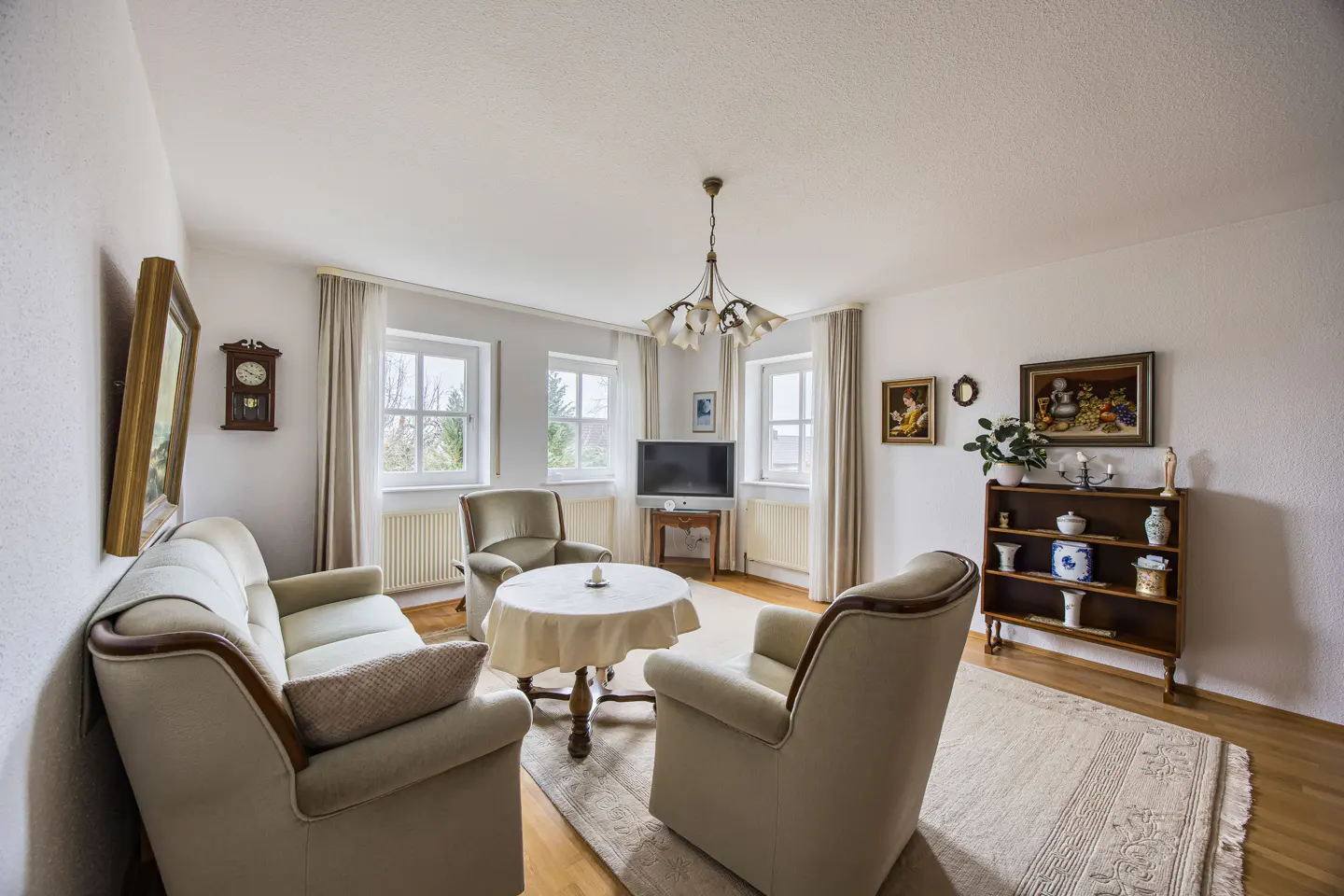Bright living room with beige sofa and chairs, round table with white cloth, TV, and wooden bookshelf on a light rug.